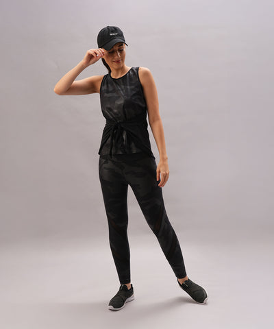 A woman in the Metallic Tie-Up - Top Only, featuring an adjustable fit and shiny finish, smiles with one hand on her cap against a plain background.