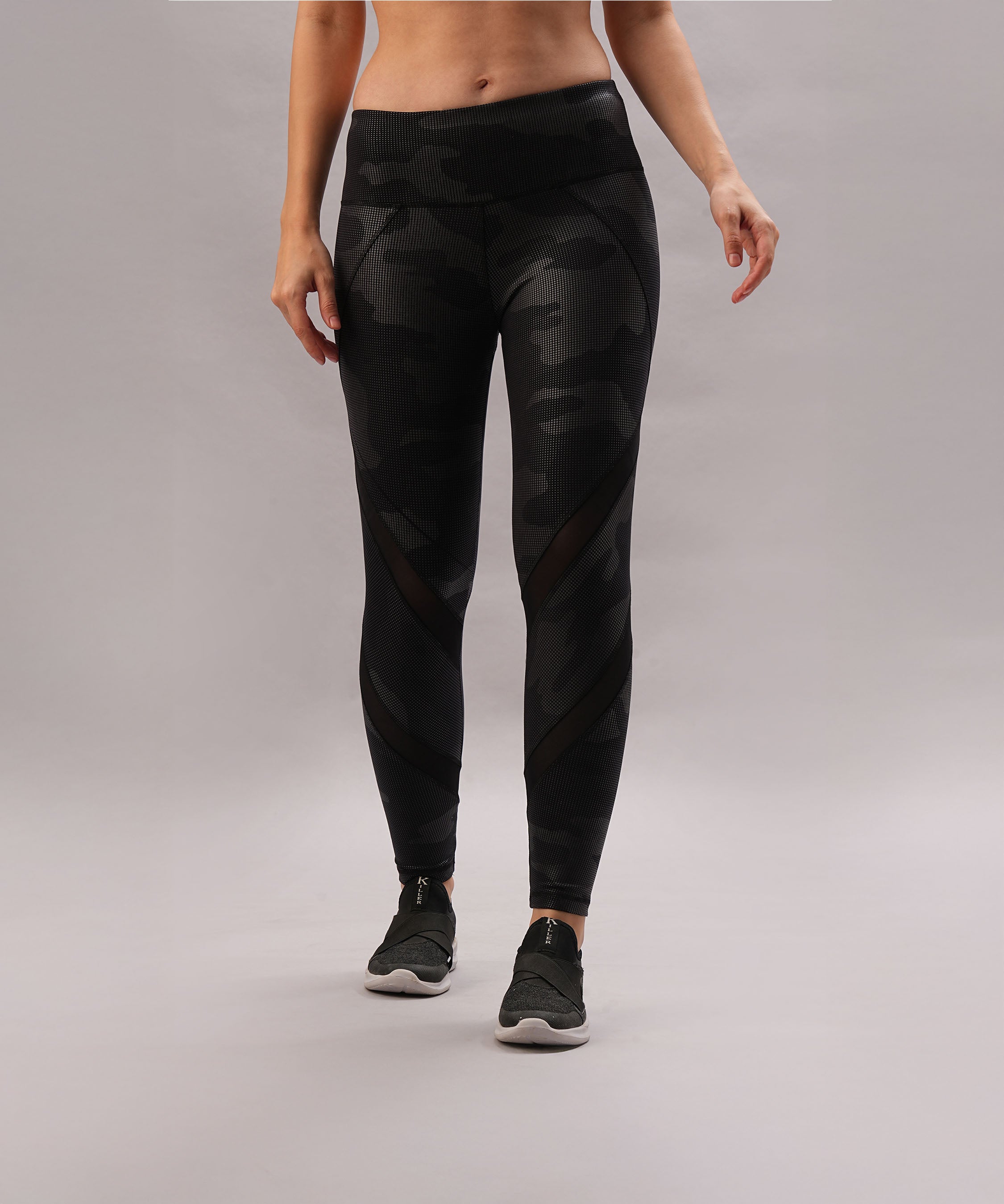 A woman stands against a plain gray background, wearing Metallic Leggings with a reflective finish and black sneakers—making a stylish statement piece for any modern wardrobe.