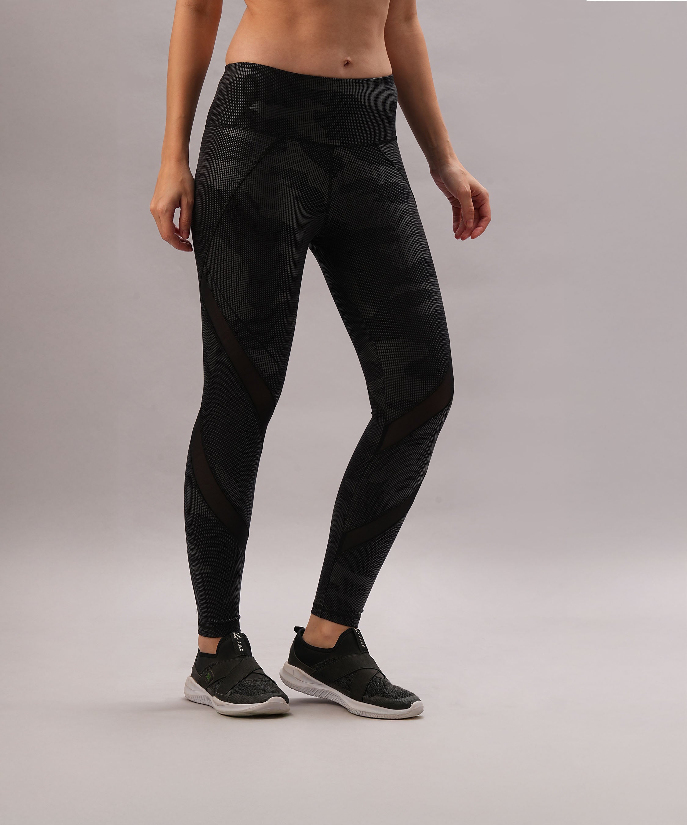 A woman stands against a plain gray background, wearing Metallic Leggings in black camo with a subtle reflective finish and black sneakers.