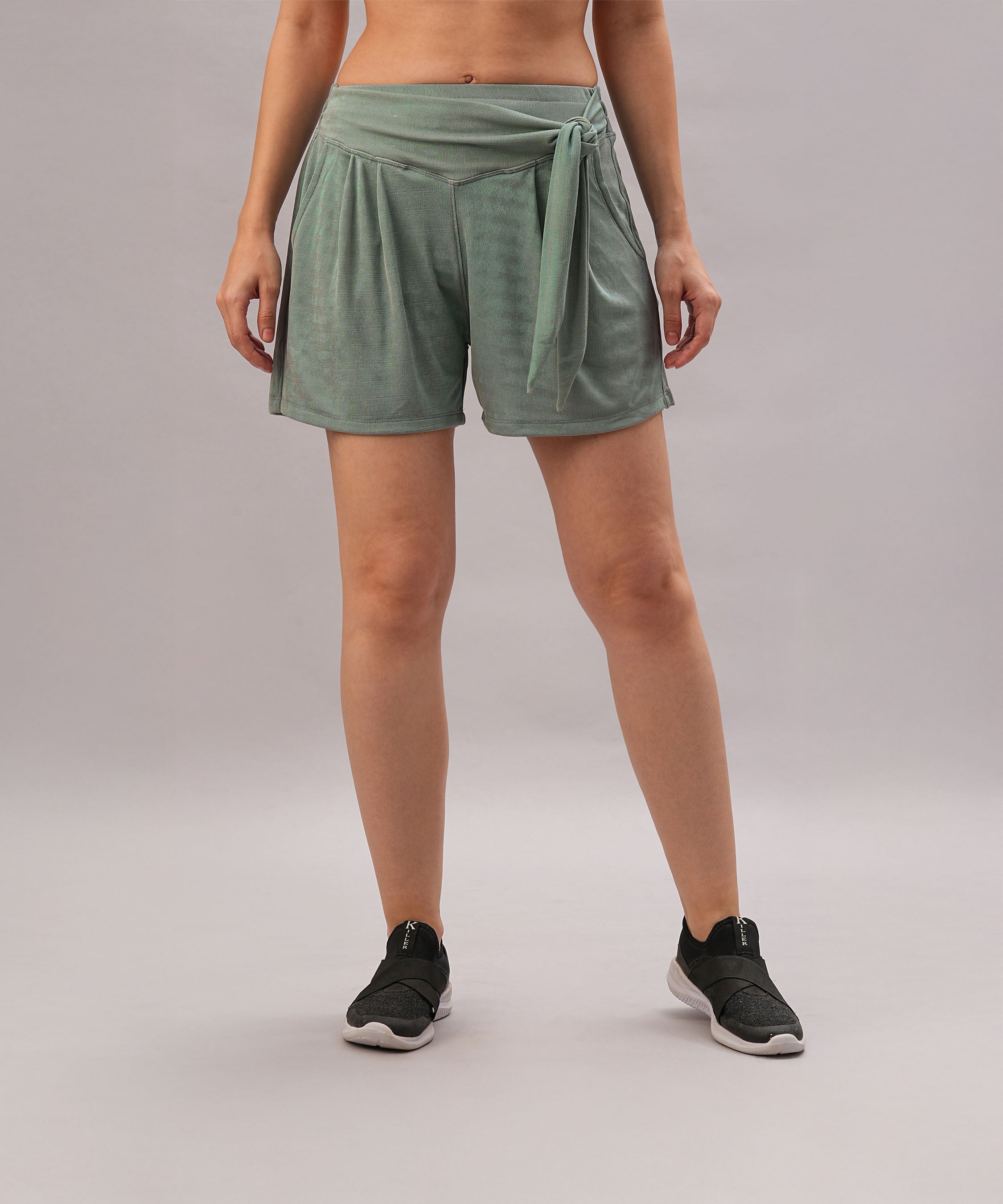 A woman models Tie up Shorts—versatile green shorts with an elastic waistband and side tie—paired with black slip-on sneakers, standing against a plain background.
