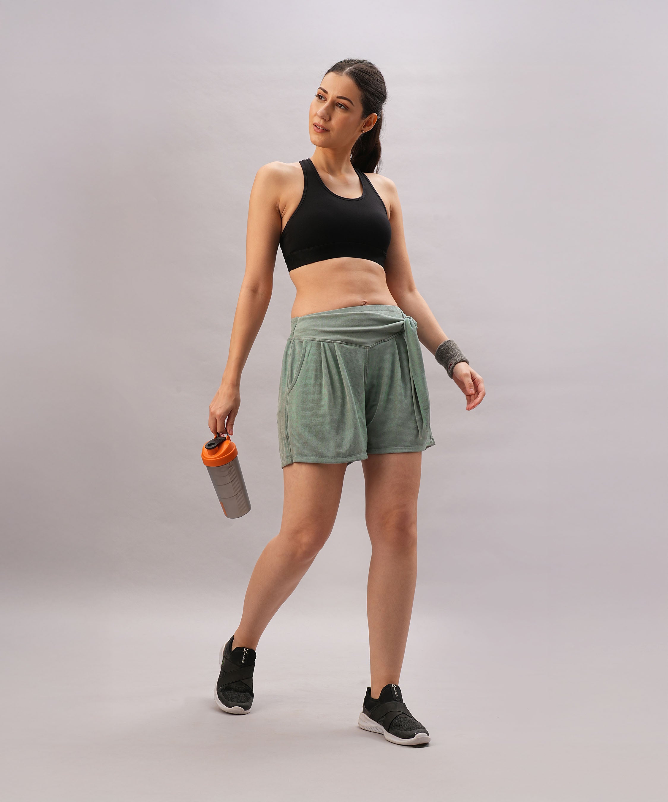 Woman in workout attire wears Tie up Shorts in metallic teal with an elastic waistband, holding a shaker bottle and standing against a plain background.