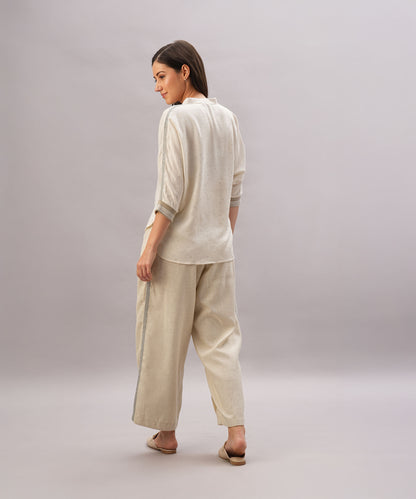 A woman in the Beige Linen Set stands barefoot, looking over her shoulder. This relaxed-fit ensemble, crafted from breathable linen, ensures effortless comfort and style.