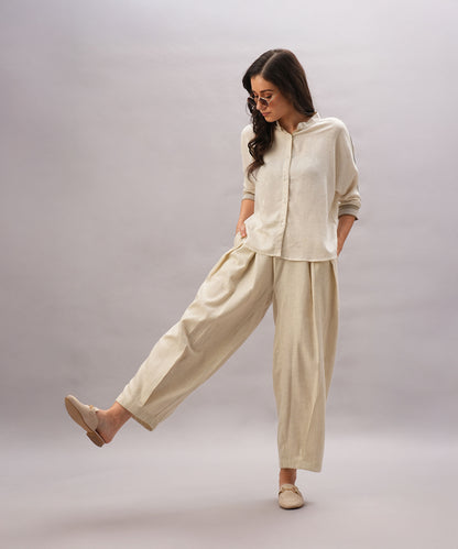A woman models the Beige Linen Set, featuring a relaxed fit and breathable linen fabric, paired with sunglasses and loafers. She poses against a plain background with one leg slightly lifted, highlighting the set’s comfort and style.