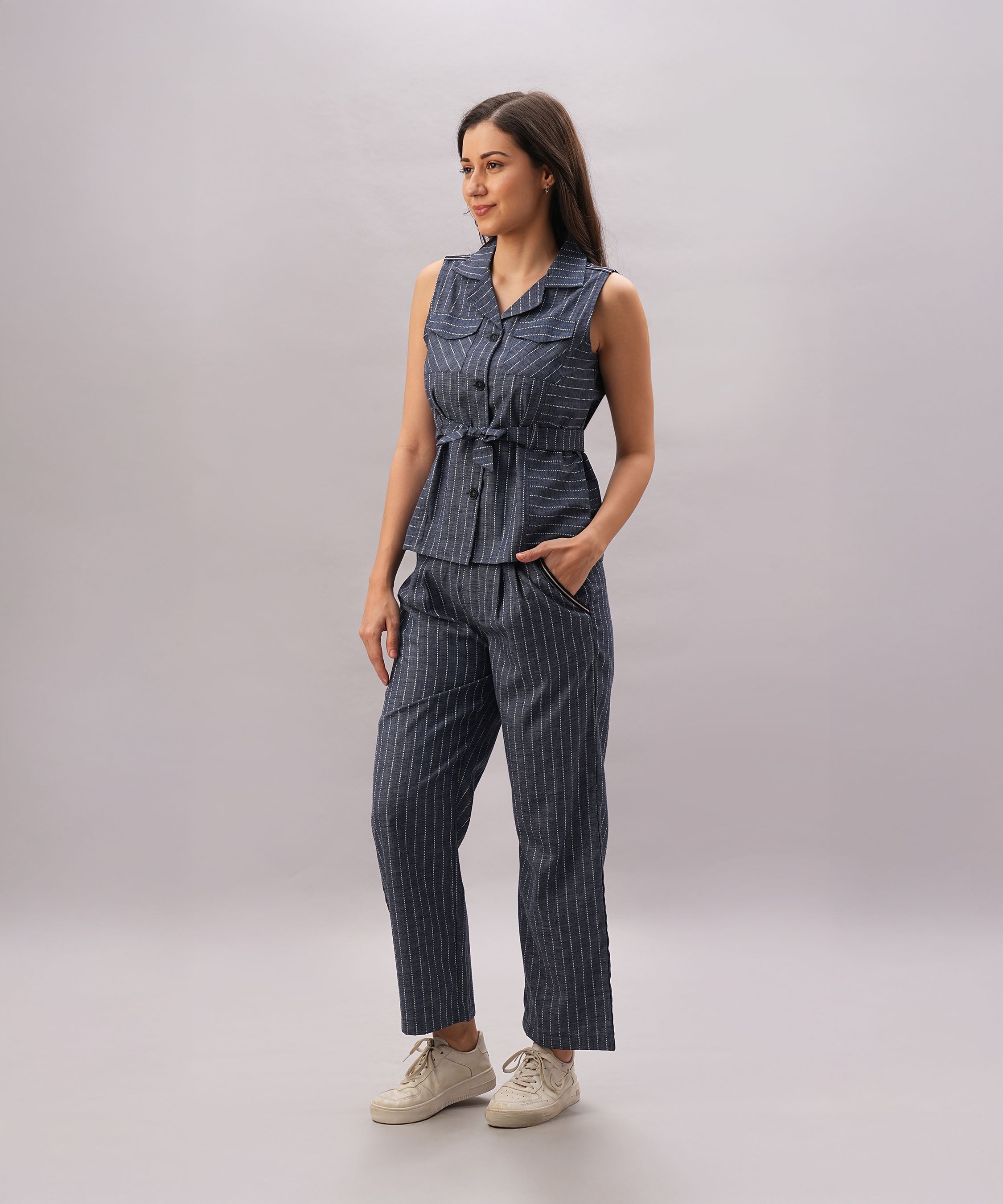 A woman wears the Navy Blue Stripe Set—a sleeveless vest and matching pants with hands in pockets, standing on a grey background. Made from 100% cotton, this set is designed for effortless style and a comfortable fit.
