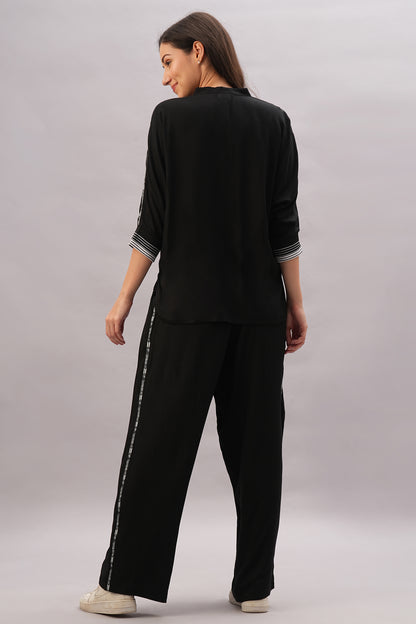 A woman stands facing away on a plain backdrop, wearing the Black Rayon Co-Ord Set featuring loose-fitting pants and a stylish top, paired with white sneakers.