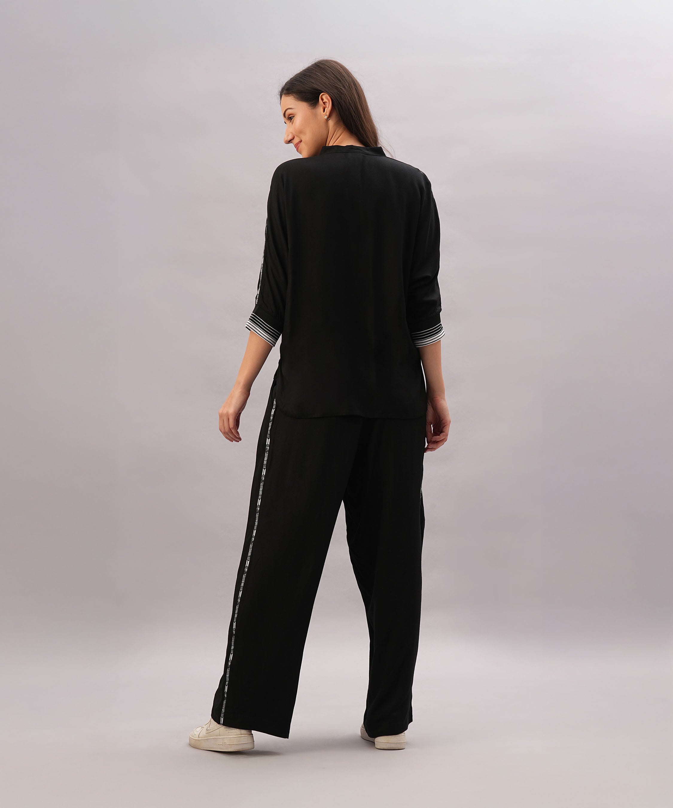 A woman faces backward against a plain light background, wearing the Black Rayon Co-Ord Set featuring a stylish top and loose-fitting pants, paired with white sneakers.