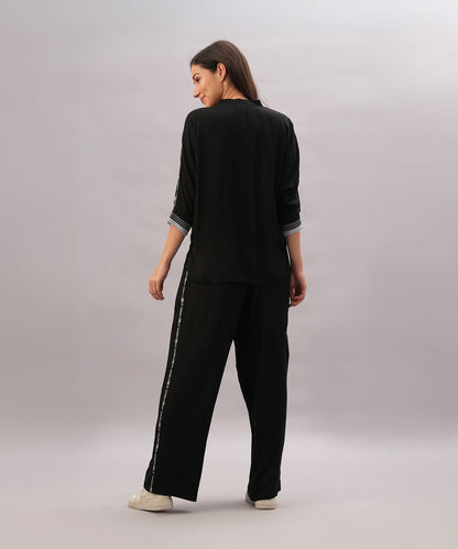 A woman faces backward against a plain light background, wearing the Black Rayon Co-Ord Set featuring a stylish top and loose-fitting pants, paired with white sneakers.