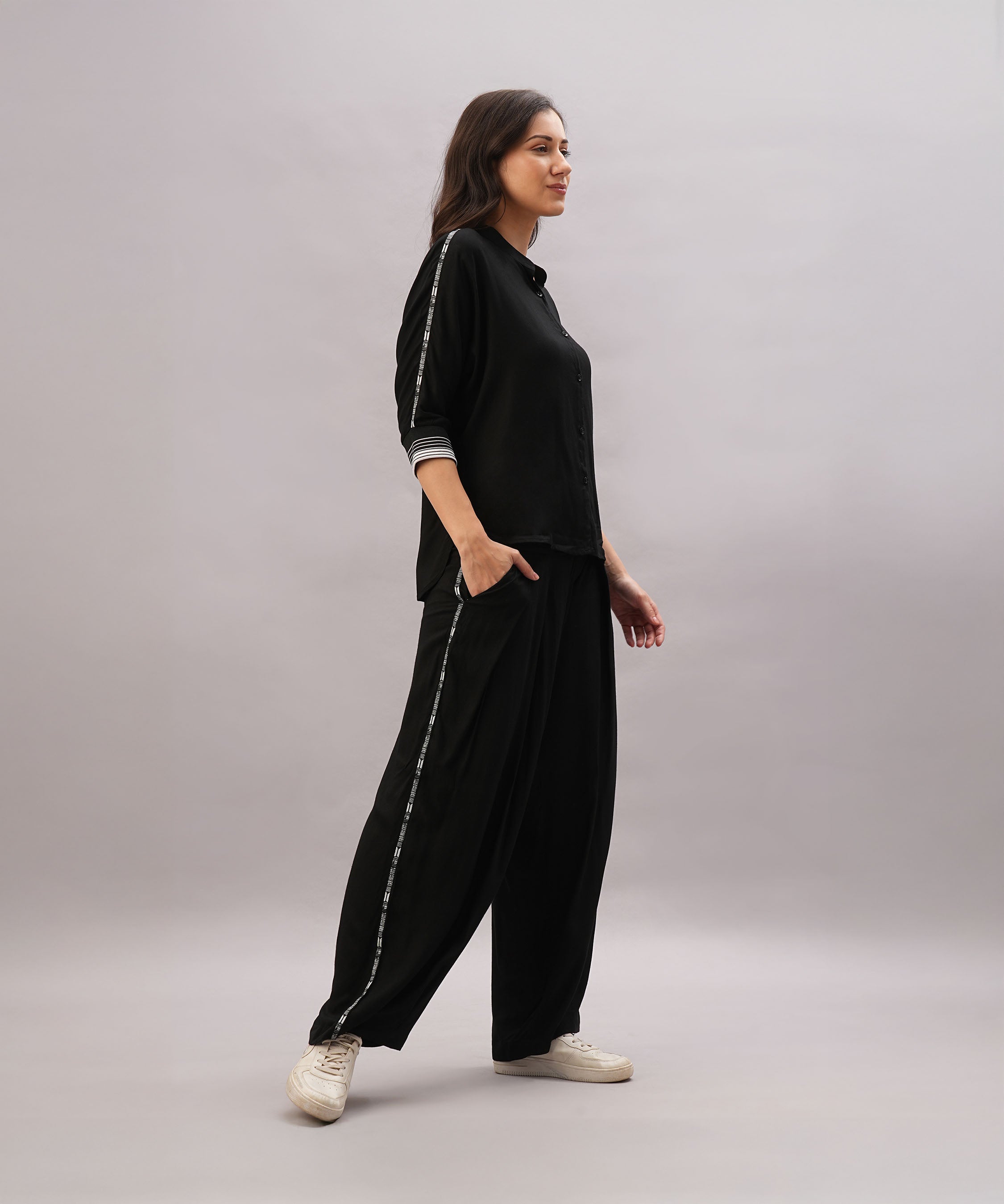 Woman wearing the Black Rayon Co-Ord Set, featuring a trendy top and wide-leg pants with side stripes, styled with white sneakers, standing sideways against a plain background.