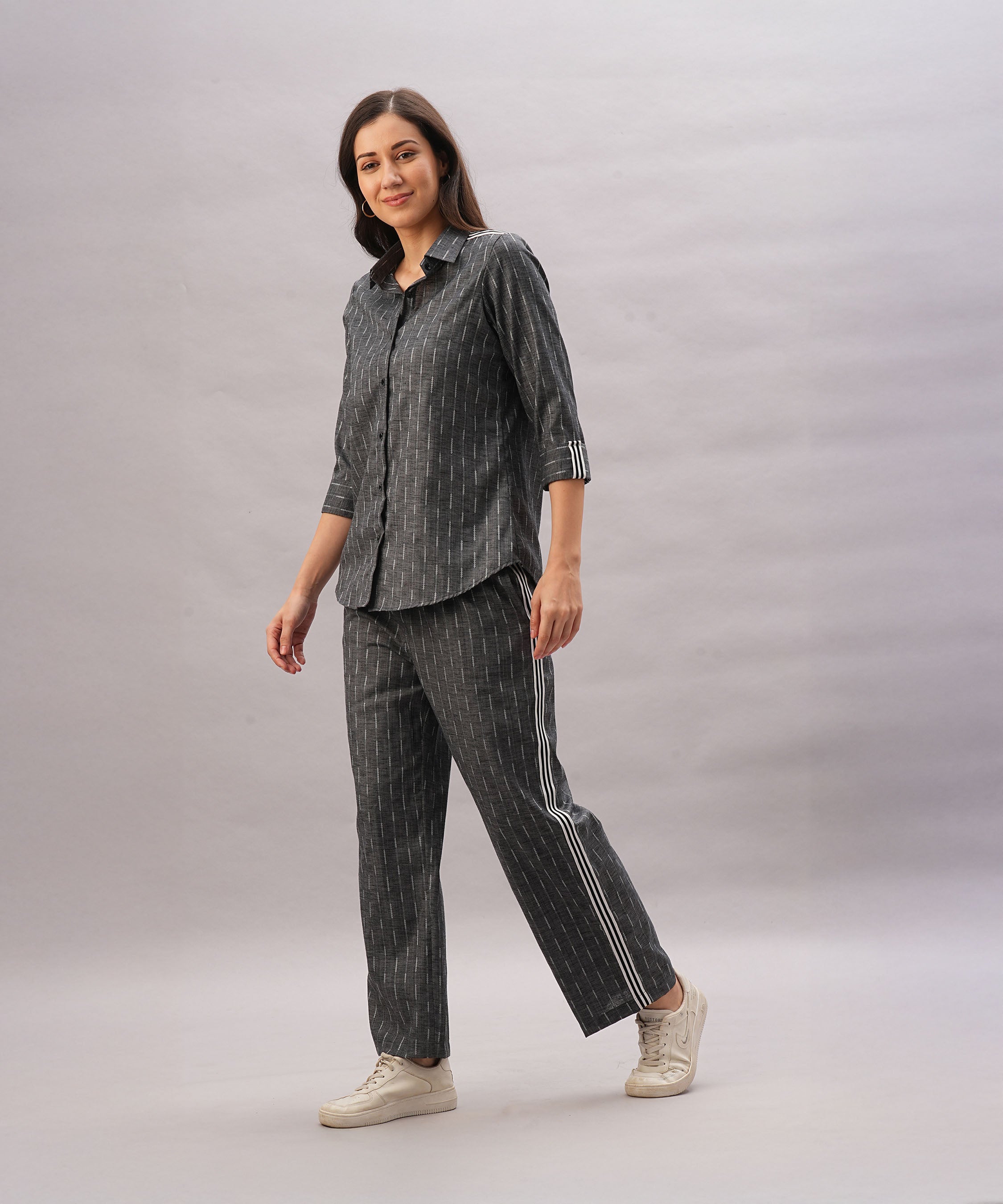 Woman in the Grey Stripes Sleeves Set—pinstripe shirt and pants styled with white sneakers—walks and smiles against a plain background, creating a perfectly coordinated look.