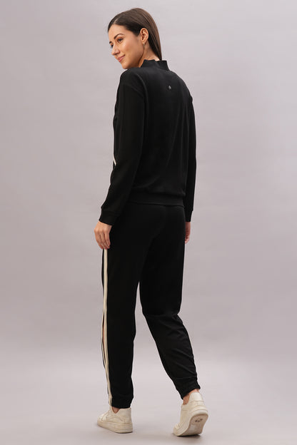 Woman wearing the Black Co-Ord Set with white side stripes, standing and looking over her shoulder against a grey background.