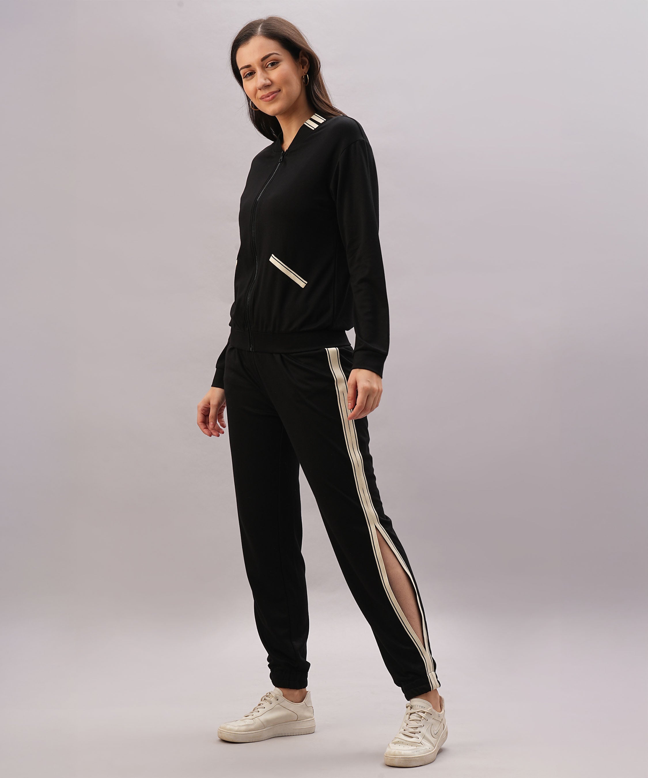 A woman stands smiling against a gray background wearing a Black Co-Ord Set with white stripes, made from cotton lycra, paired with white sneakers.