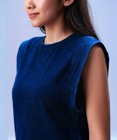 A woman wears the Blue Denim Wash Capsleeves Top, standing against a light blue background and facing slightly sideways.