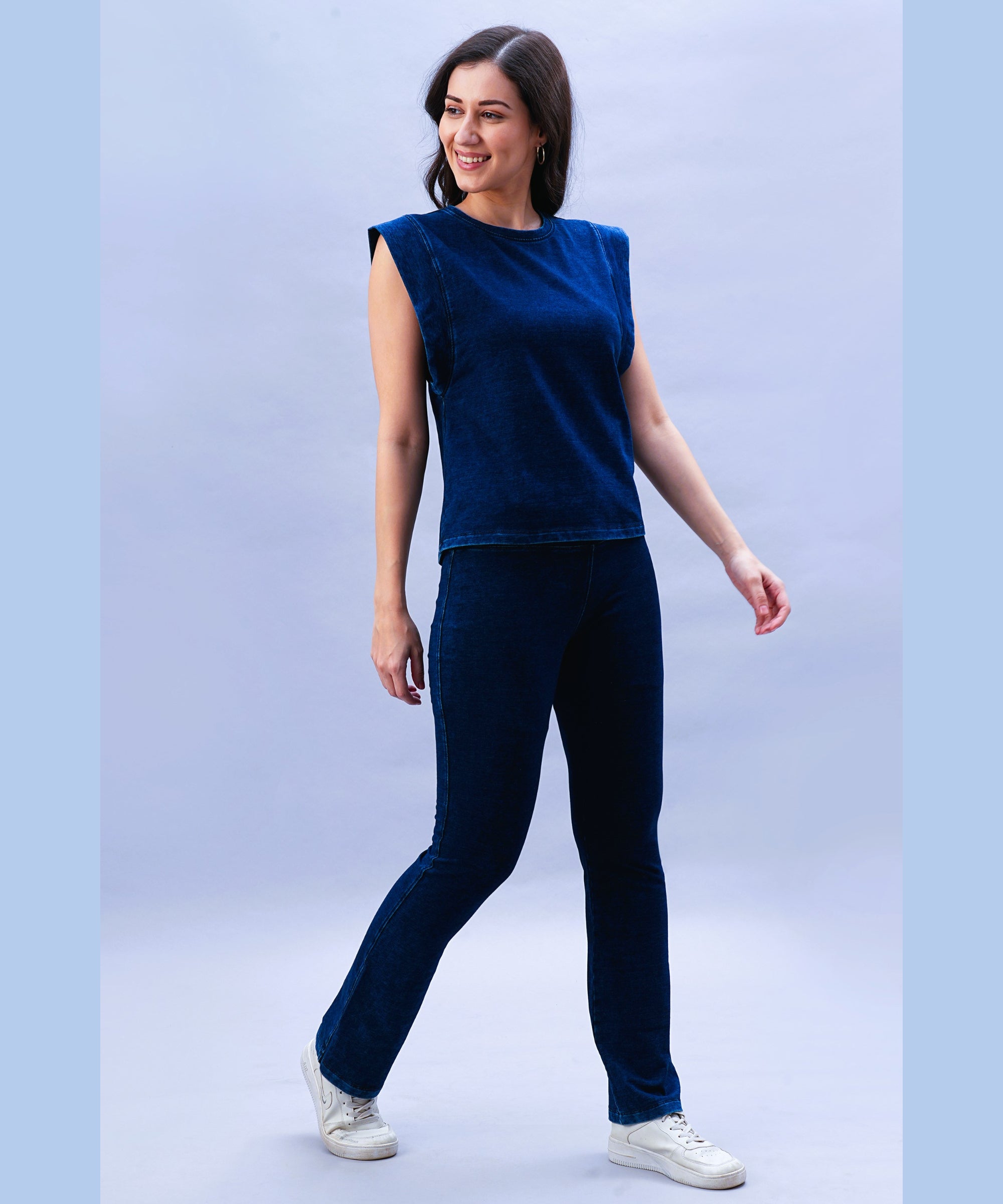 Woman stands smiling against a light background, wearing Blue Denim Wash Bootcut Leggings with a high-rise waistband, sleeveless navy blue top, and white sneakers.