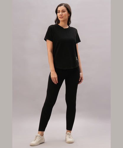 Woman wearing the Black Denim Wash Workout Tee and black leggings with white sneakers, standing against a plain background—offers style and moisture-wicking comfort, ideal for athletic performance.