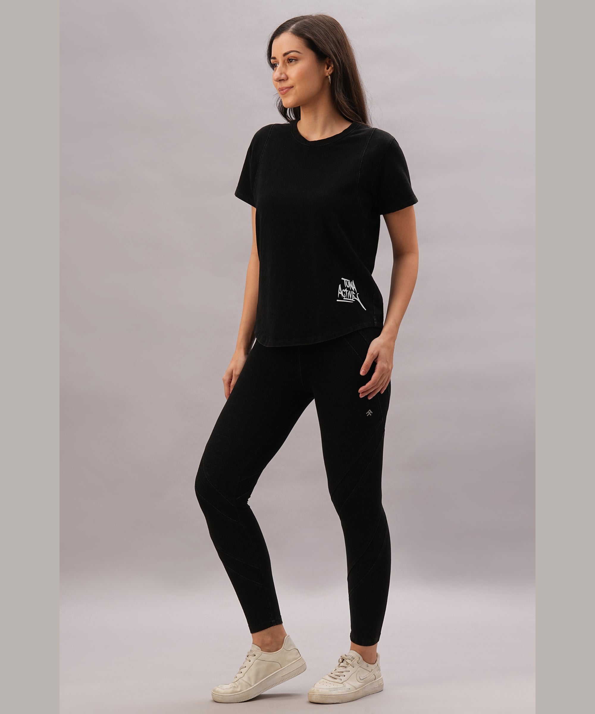 A woman poses against a plain background wearing a black t-shirt and the Black Denim Wash Leggings, featuring a high-rise waistband and four-way stretch fabric, paired with white sneakers.