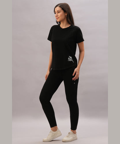 A woman poses against a plain background wearing a black t-shirt and the Black Denim Wash Leggings, featuring a high-rise waistband and four-way stretch fabric, paired with white sneakers.