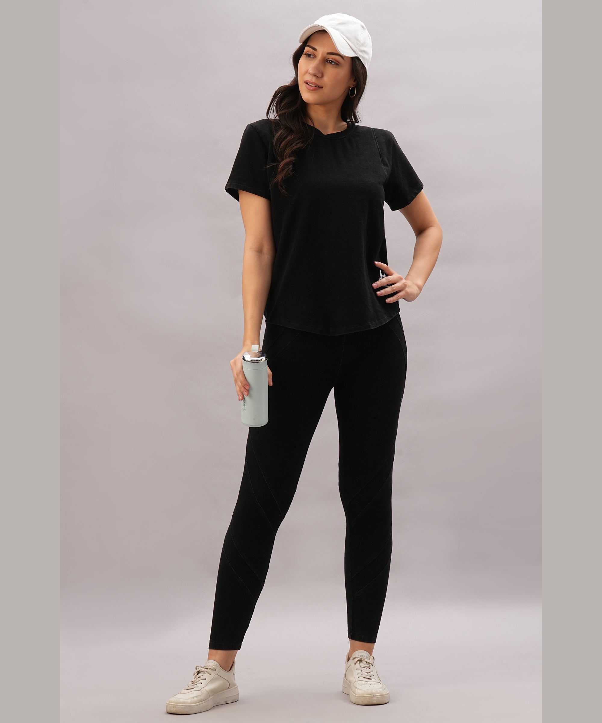 A woman in a black athletic outfit and white cap holds a water bottle, confidently wearing the Black Denim Wash Workout Tee for optimal sweat-wicking performance.