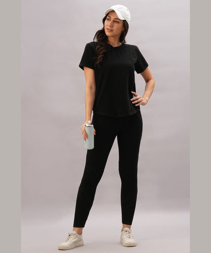 A woman in a black athletic outfit and white cap holds a water bottle, confidently wearing the Black Denim Wash Workout Tee for optimal sweat-wicking performance.