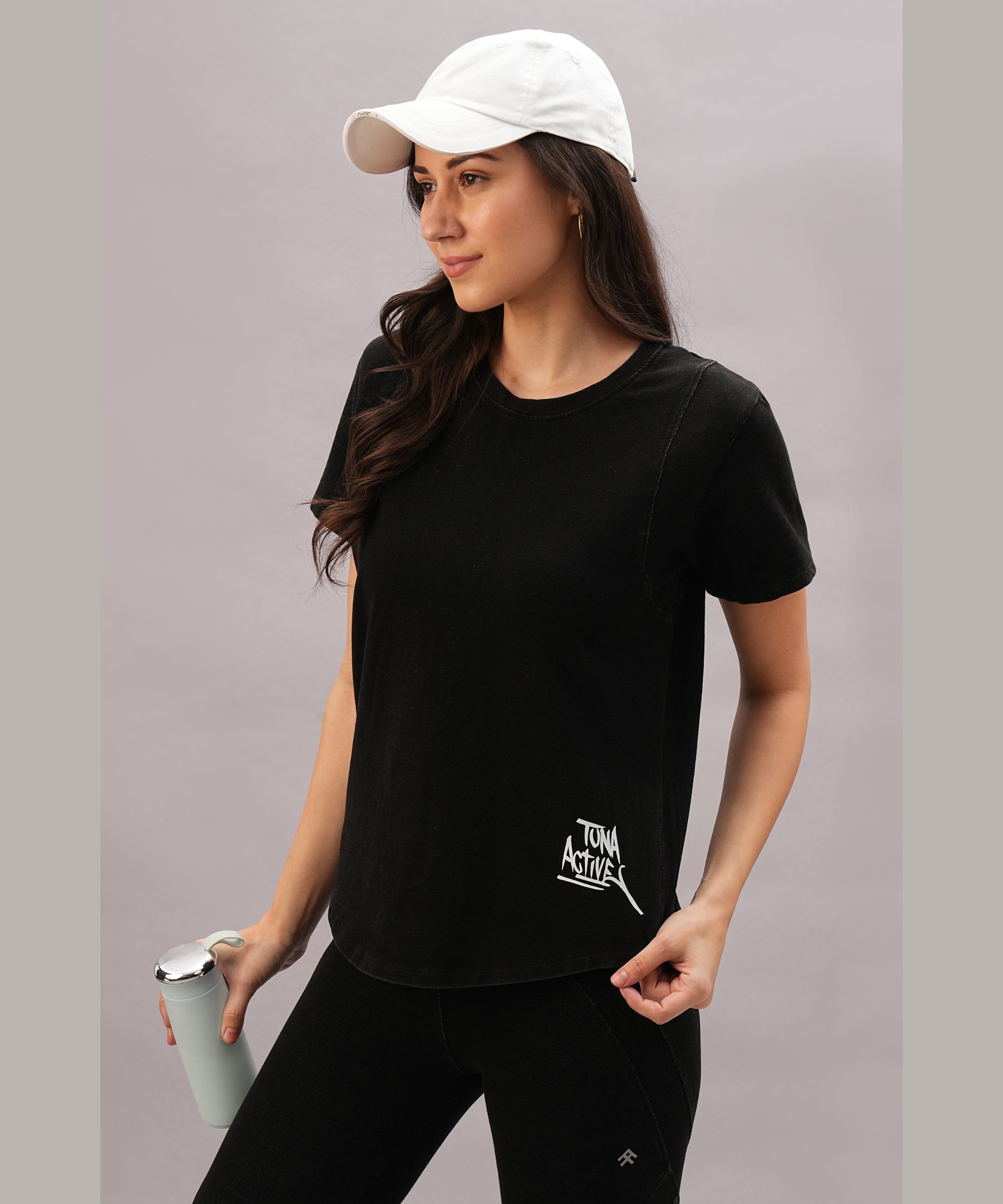 Woman in a Black Denim Wash Workout Tee and white cap holds a tumbler against a light background—ideal for boosting athletic performance with this stylish, moisture-wicking tee.