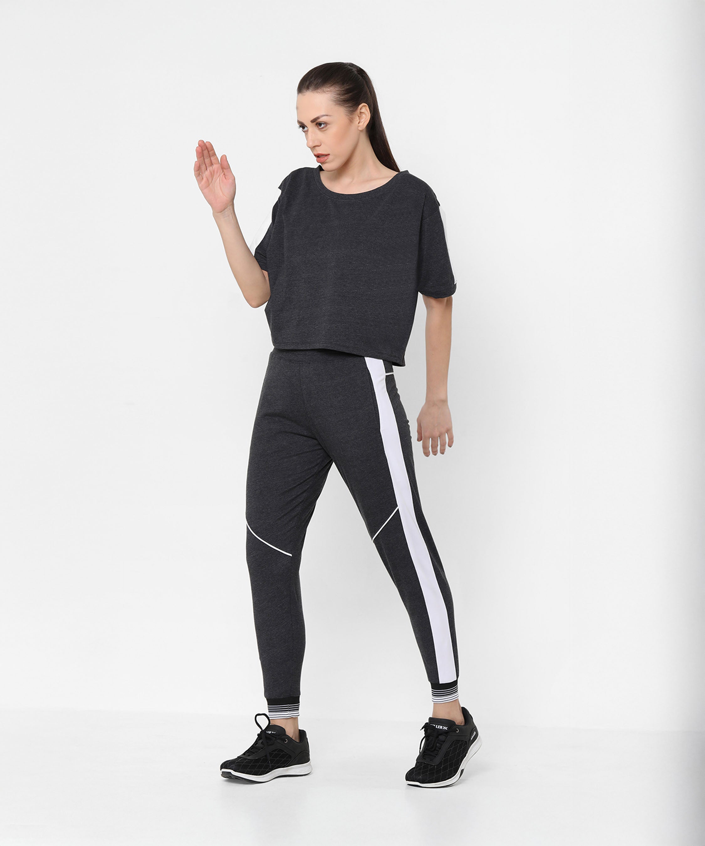 A woman models the Melange Dark Grey Set, featuring dark gray joggers with white side stripes and a comfy fit top, against a plain white background.