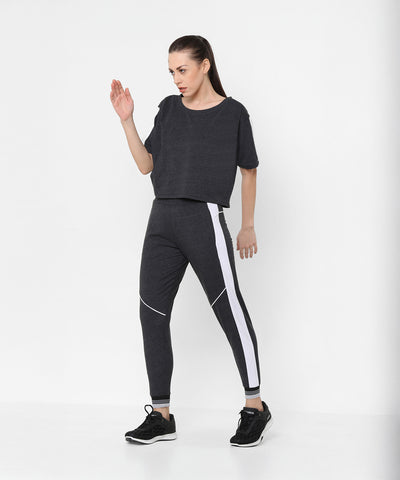 A woman models the Melange Dark Grey Set, featuring dark gray joggers with white side stripes and a comfy fit top, against a plain white background.