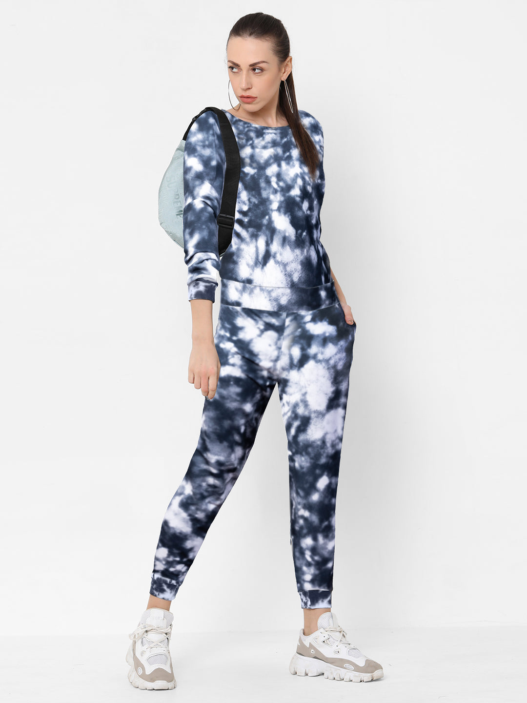 Woman in a black and white Tie and Dye Tracksuit Set with white sneakers and a light blue backpack stands against a white background—an effortless, stylish look that elevates your fashion game.