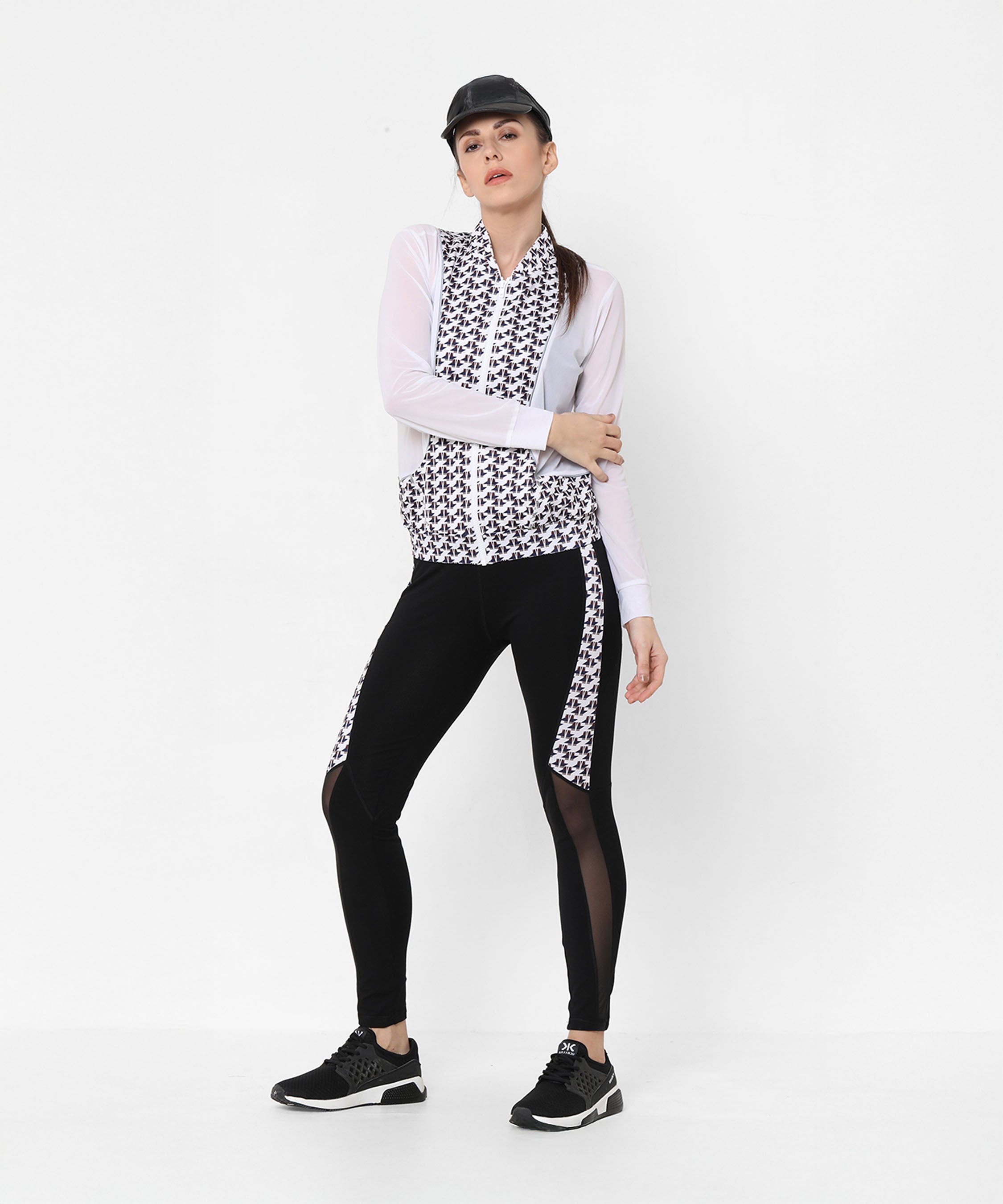 Woman exudes confidence in the Esther Jacket With Leggings set, pairing a printed net jacket with black leggings and completing the athleisure look with a cap and black sneakers against a white background.