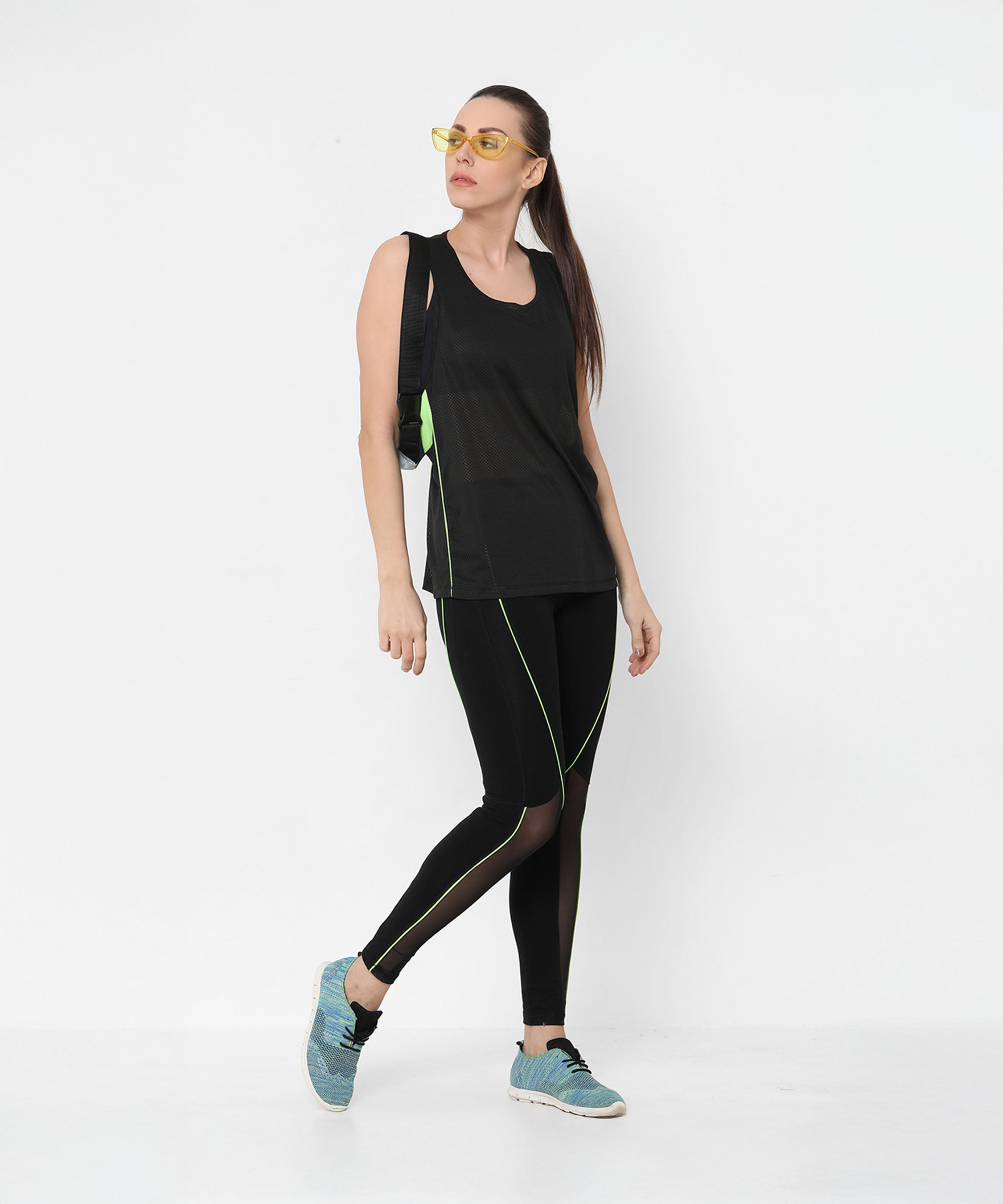 A woman poses against a plain white background wearing Fab Fit Neon Pop Leggings with a high-rise waistband, yellow sunglasses, and blue shoes.