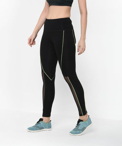 A woman wears the Fab Fit Neon Pop Leggings featuring black mesh, green piping, and a high-rise waistband, paired with blue sneakers and a dark crop top.