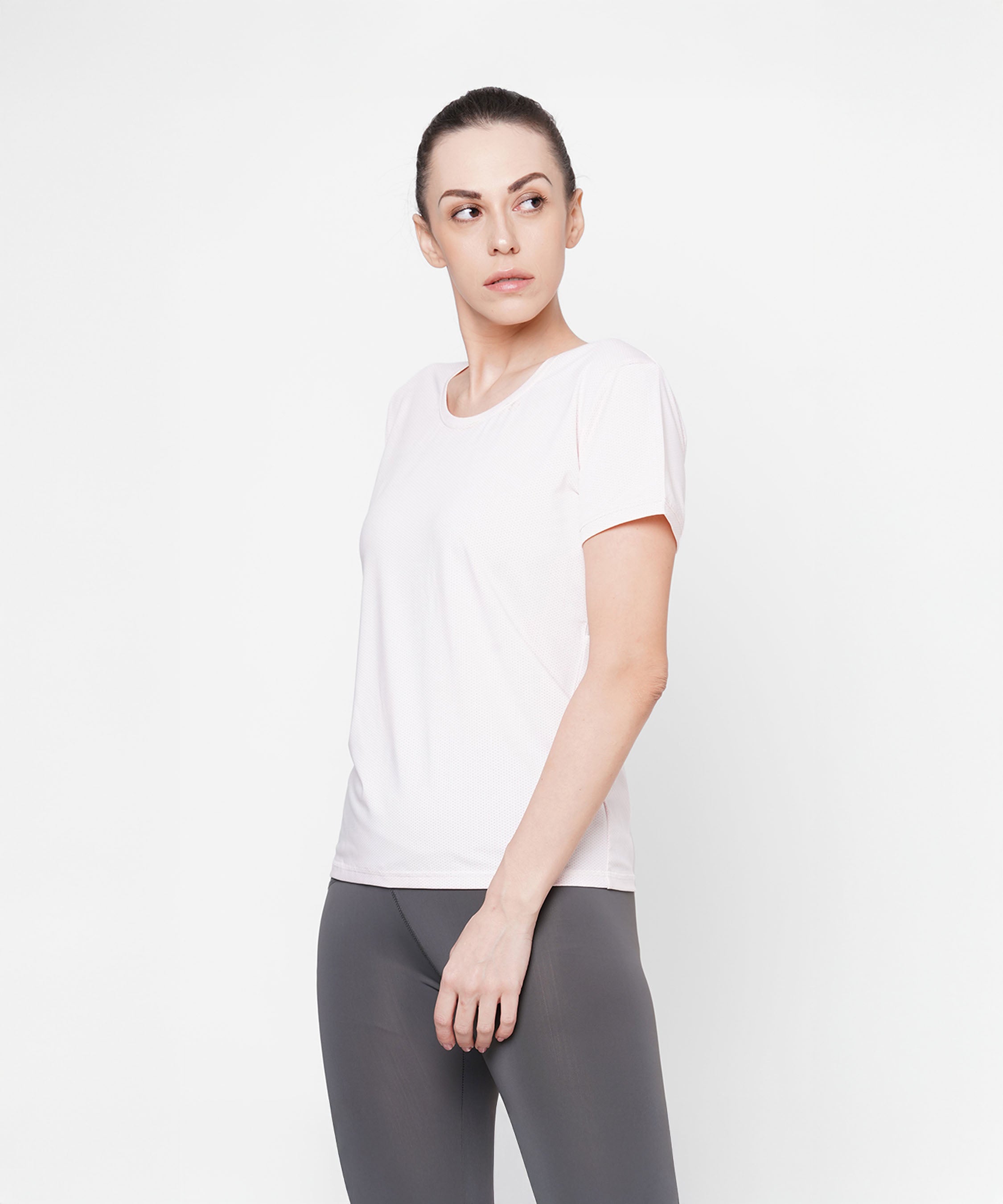 Woman wearing a light pink round neck t-shirt and gray leggings stands against a white background, looking to the side.