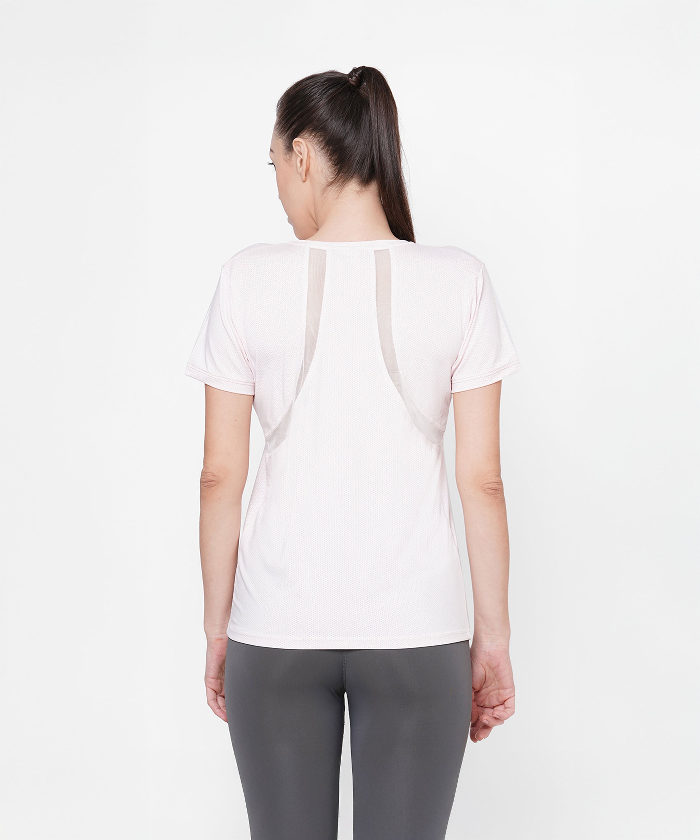 A woman stands with her back to the camera, wearing a Light Pink Round Neck T-Shirt and gray leggings against a white background.