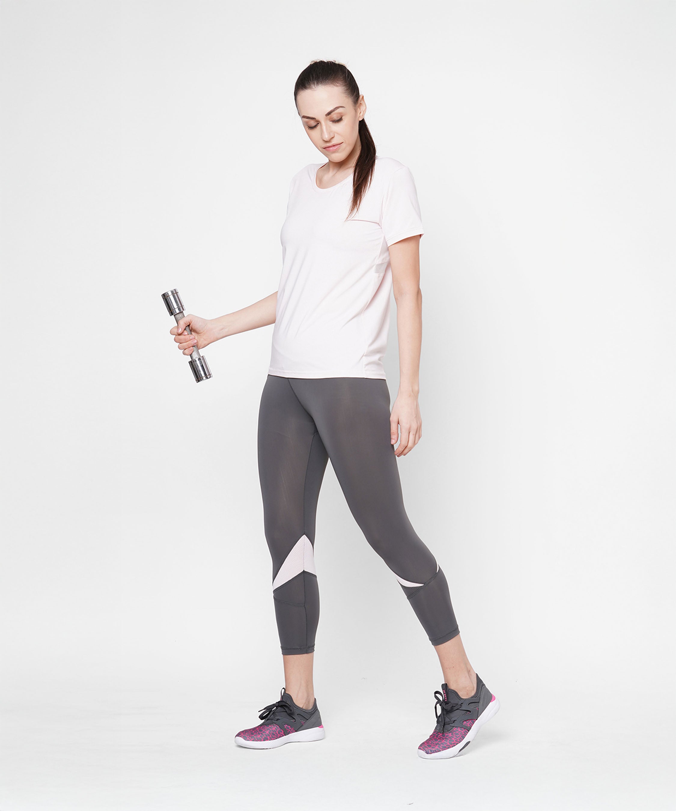 A woman in a light pink round neck T-shirt and leggings holds a dumbbell, standing confidently against a plain white background.