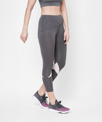 Woman wearing Classic Capri grey leggings with a thick waistband, paired with a gray sports bra and gray sneakers with pink accents, standing against a white background.