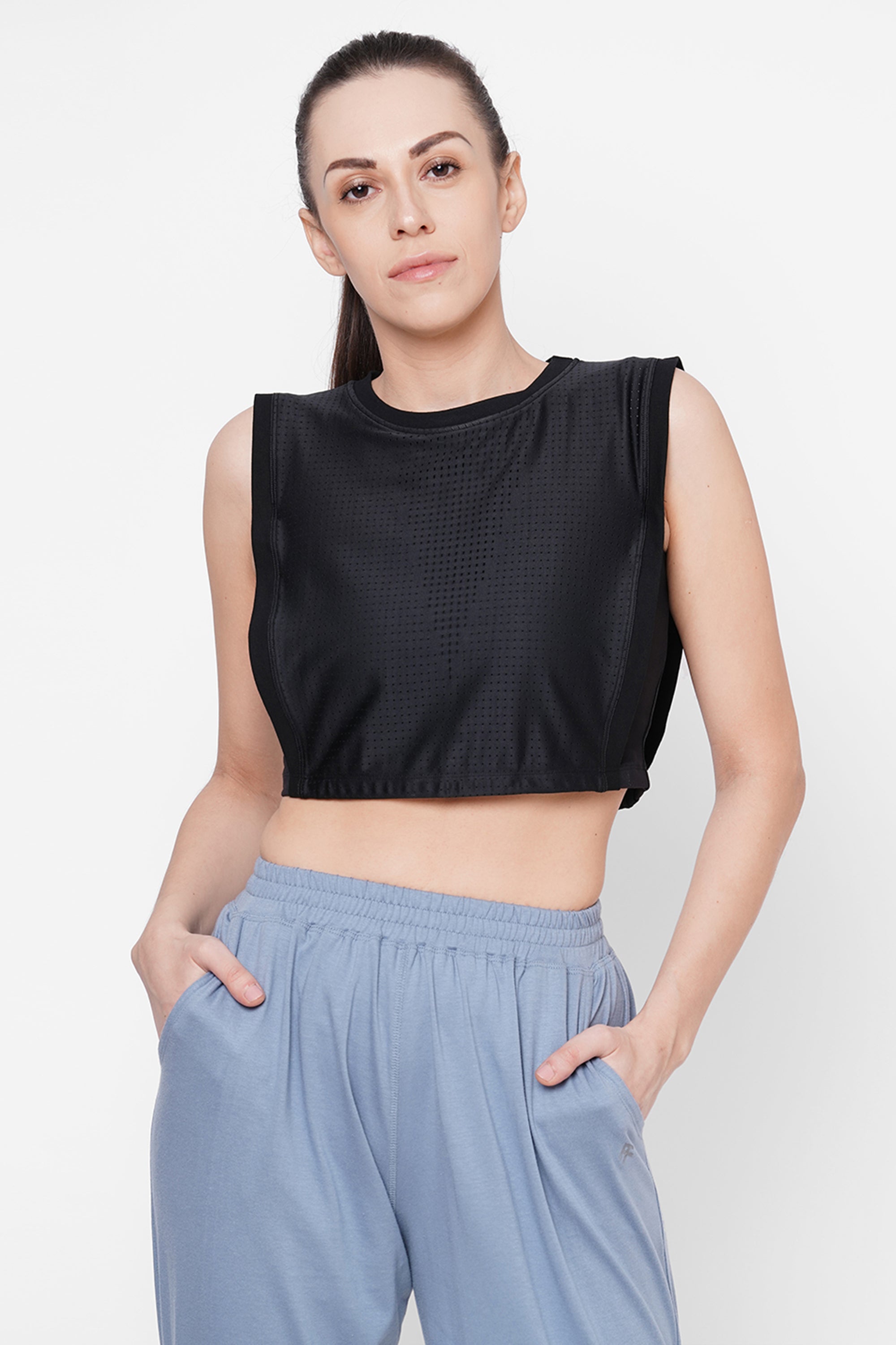 Woman wearing a Black Crop Top With Mesh Overlay and light blue pants, hands in pockets, standing against a white background.