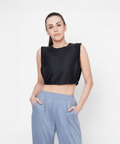 Woman in a Black Crop Top With Mesh Overlay and light blue pants stands with hands in pockets against a white background.