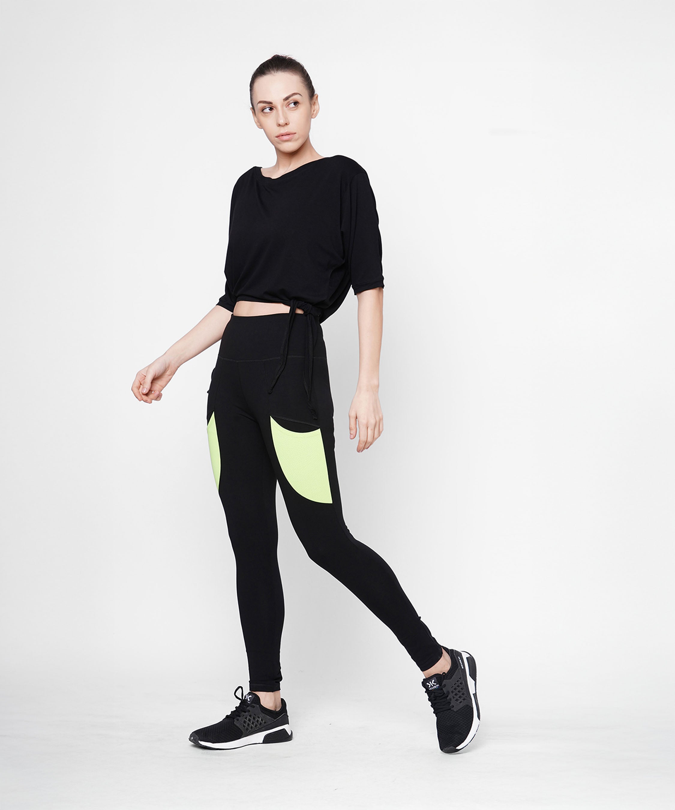 A woman poses against a plain white background wearing Black Leggings With Neon Green Patch, featuring a high-rise waistband and neon green patch pockets.