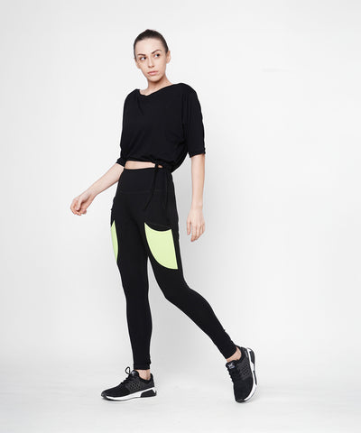 A woman poses against a plain white background wearing Black Leggings With Neon Green Patch, featuring a high-rise waistband and neon green patch pockets.