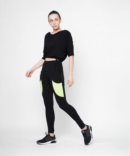 A woman poses against a plain white background wearing Black Leggings With Neon Green Patch, featuring a high-rise waistband and neon green patch pockets.