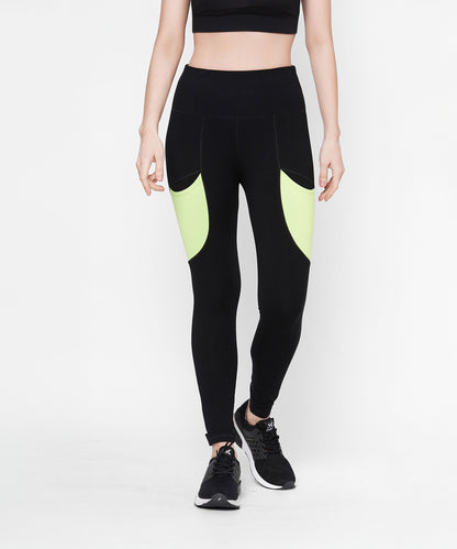 A woman is wearing Black Leggings With Neon Green Patch, featuring side panels and a high-rise waistband, paired with a black crop top and black running shoes.