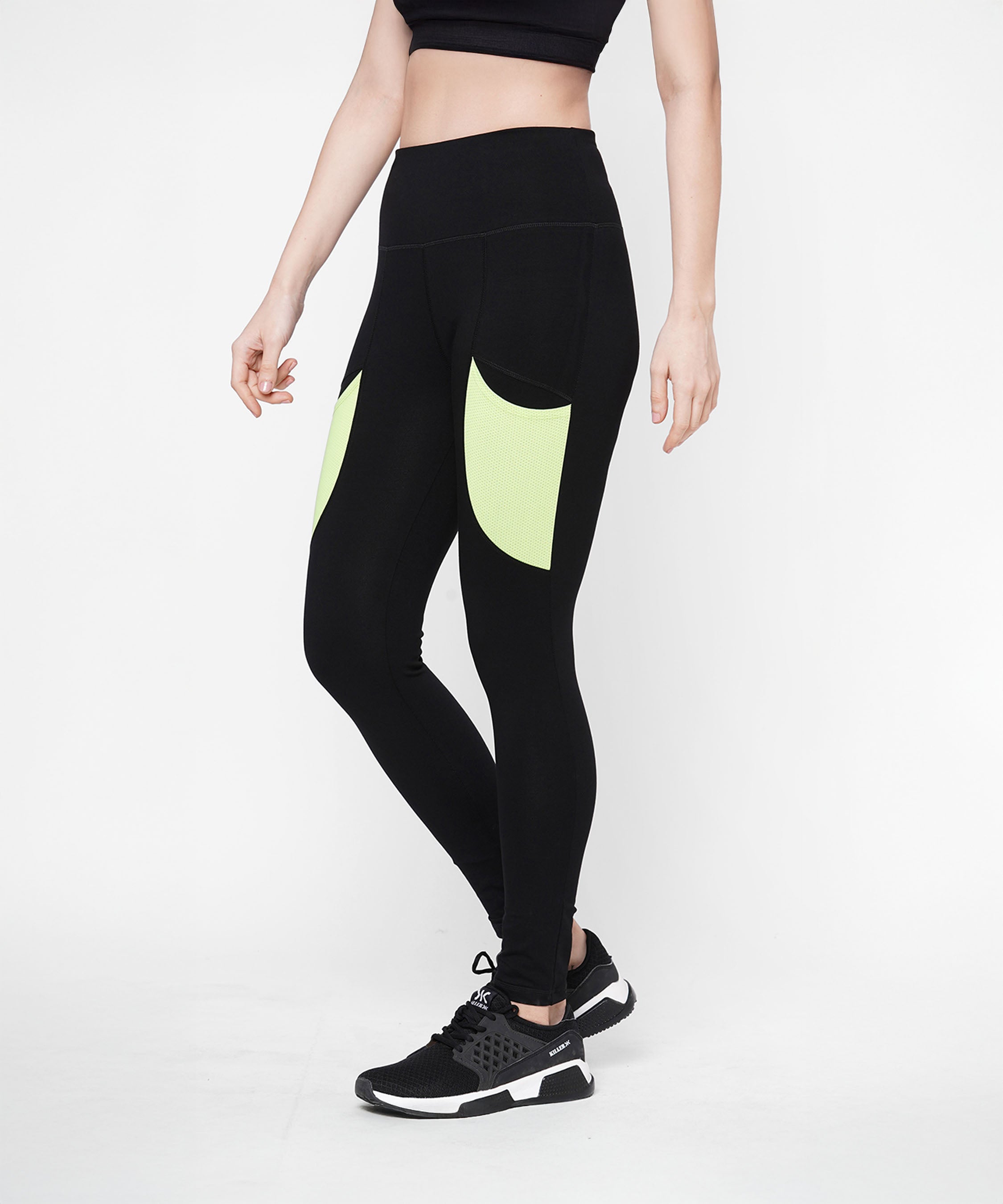 A woman in Black Leggings With Neon Green Patch, featuring a high-rise waistband and neon green patch pockets, pairs them with a black crop top and black sneakers while standing against a white background.