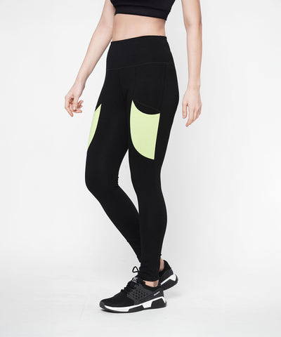 A woman in Black Leggings With Neon Green Patch, featuring a high-rise waistband and neon green patch pockets, pairs them with a black crop top and black sneakers while standing against a white background.