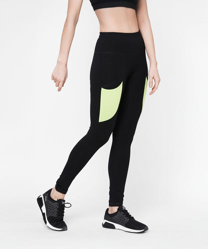 A woman wears Black Leggings With Neon Green Patch featuring a high-rise waistband and neon green patch pockets, styled with black sports shoes and a black crop top.