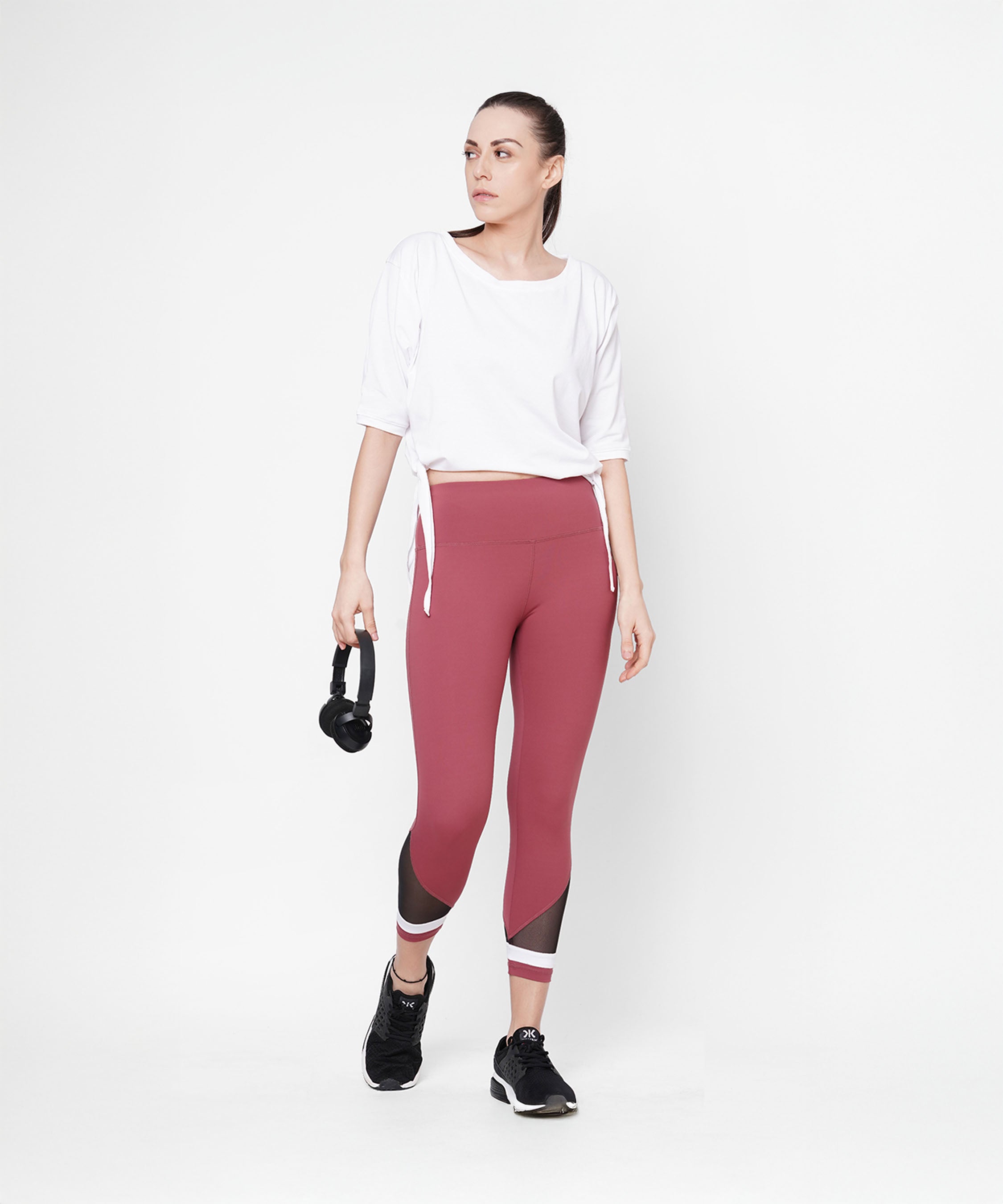 A woman in athletic wear wears Punch Pink Capri with a compression fit waistband, holding headphones against a plain white background.