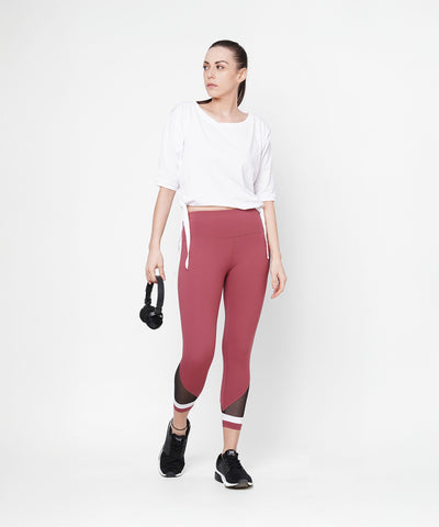 A woman in athletic wear wears Punch Pink Capri with a compression fit waistband, holding headphones against a plain white background.