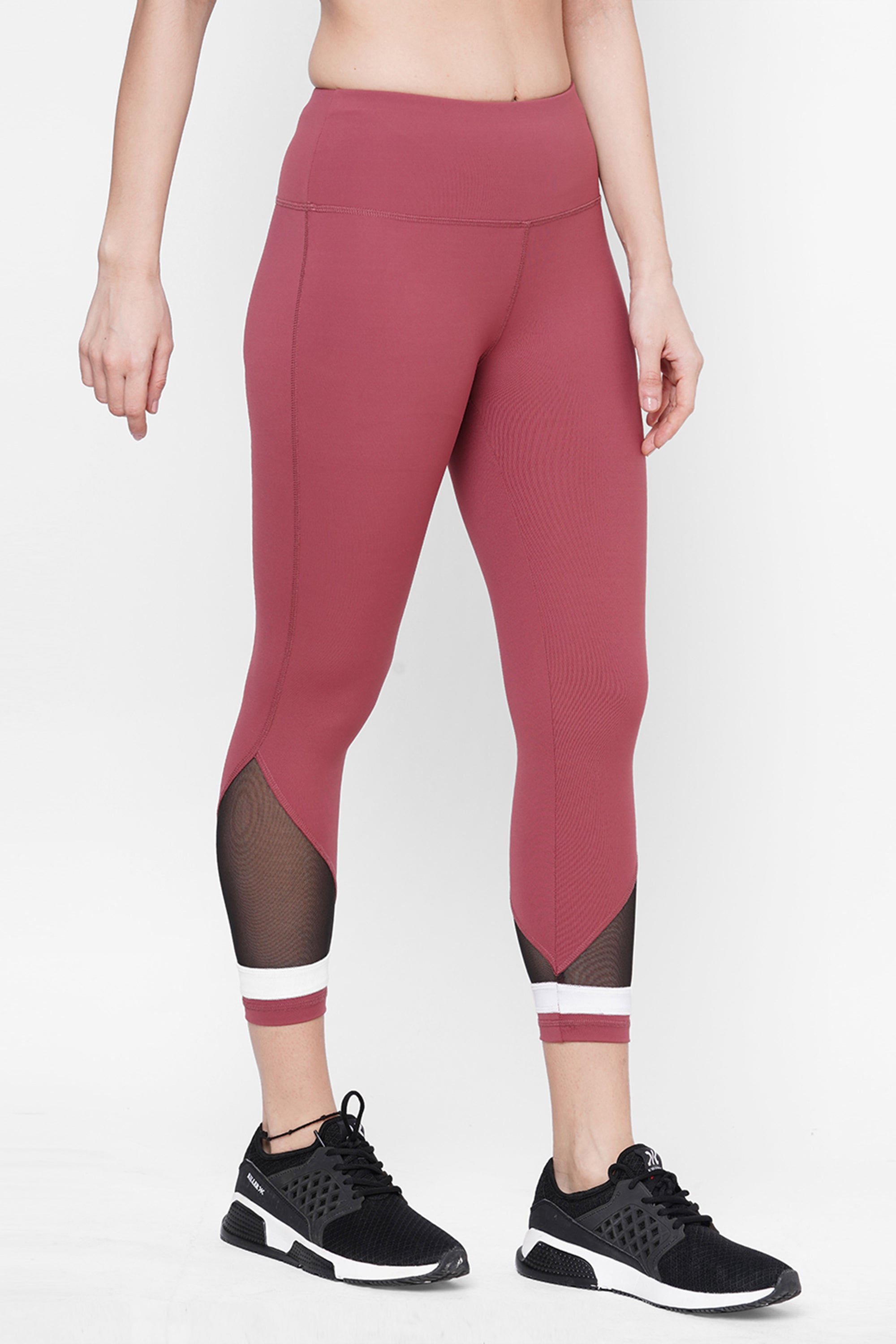 A person wears the Punch Pink Capri—high-waisted mauve workout leggings with black mesh panels and a compression fit waistband—paired with black sneakers, standing on a white background for a stylish athletic look.