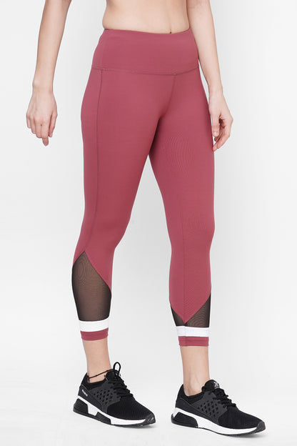 A person wears the Punch Pink Capri—high-waisted mauve workout leggings with black mesh panels and a compression fit waistband—paired with black sneakers, standing on a white background for a stylish athletic look.