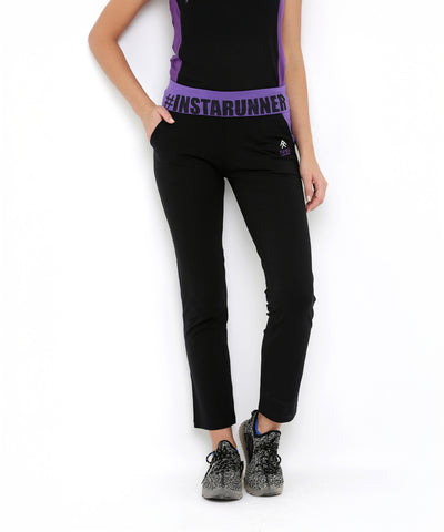 A person wearing the Regular Fit Trackpants TLC05 with a branded "#INSTARRUNNER" waistband, styled with a black top featuring purple accents and black sneakers.