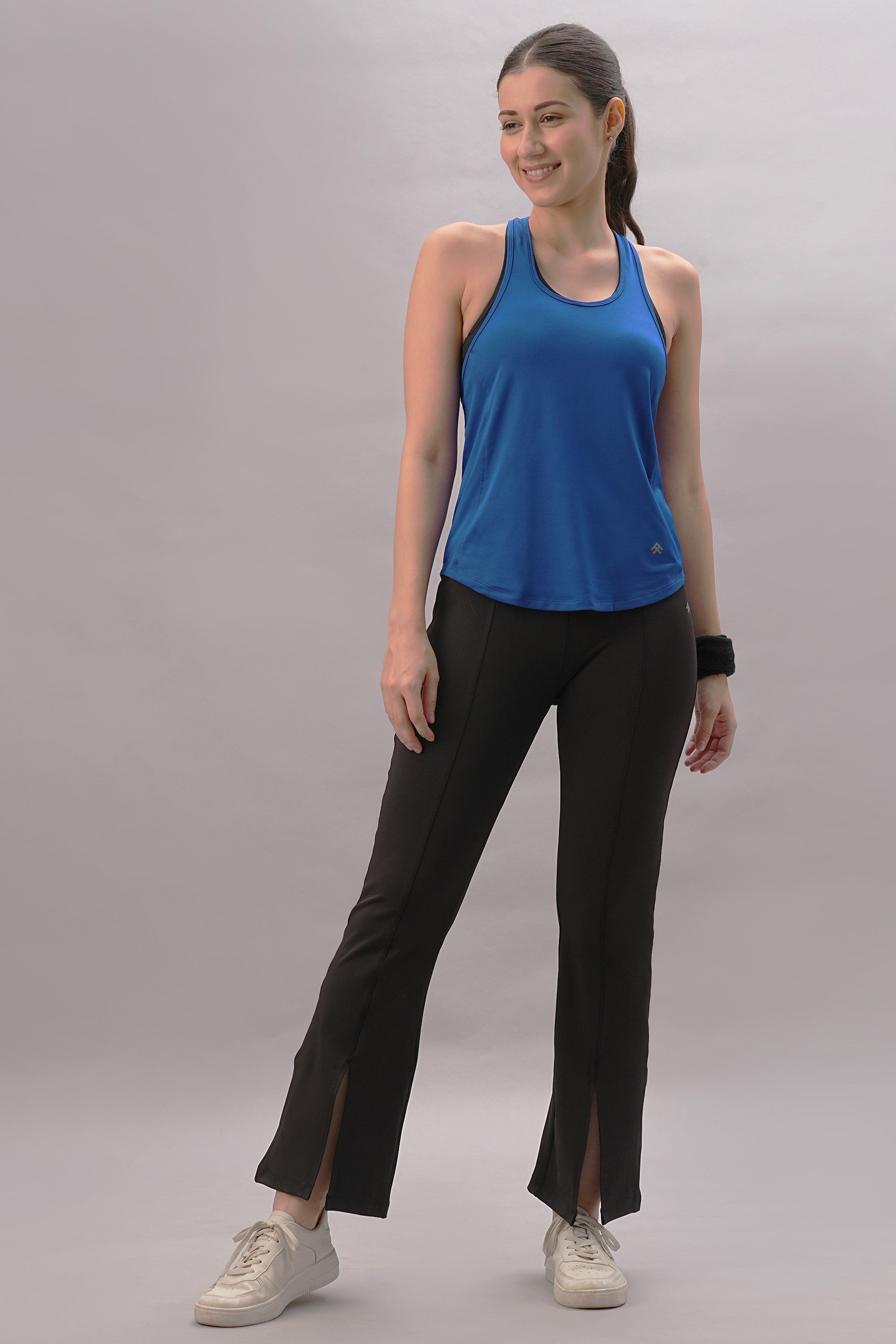 Woman in a sporty, stylish look, wearing the Blue Racer Back Top, black pants with slits, and white sneakers, standing and smiling.