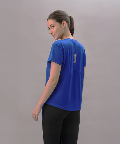 Woman wearing the Blue Comfort T-Shirt and black pants stands sideways, glancing over her shoulder against a light background—a stylish pick for an active lifestyle.
