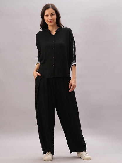 A woman wears the Black Rayon Co-Ord Set, featuring a loose black button-up top and wide-leg pants, standing with her hands in her pockets.